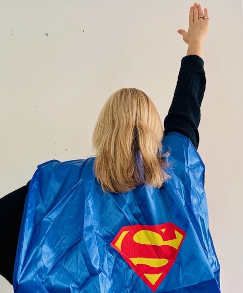 A woman wearing a superhero cape holding one arm up in the air as though she is about to start flying