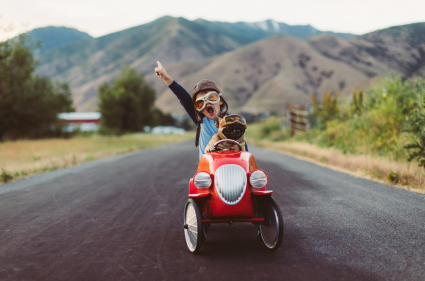Small boy and dog are driving in a small go-kart together. The boy has his hand in the air with an excited expression.